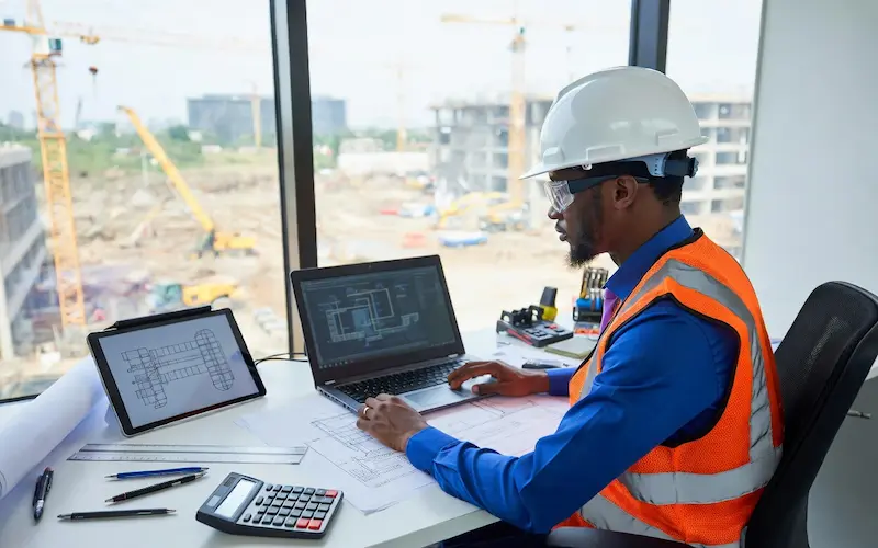 MEP Engineer at work in Kenya - reviewing HVAC, electrical, and plumbing system designs on laptop and tablet in a modern construction site office