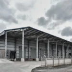 Photorealistic 3D exterior render of a large industrial warehouse in Athi River, Kenya. Features steel portal frame structure, grey metal cladding, large roller shutter doors, loading dock with concrete ramp, perimeter fence, and clear signage under overcast sky. Architectural and structural visualization by Cadreatech Engineering Services Ltd, Nairobi – experts in industrial projects across Kenya.