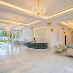 A high-end luxury beach hotel lobby in Diani, Kenya, featuring a text-free blue and gold reception desk, ornate coffers ceiling with crystal chandeliers, and expansive window arches overlooking the Indian Ocean, by Cadreatech.